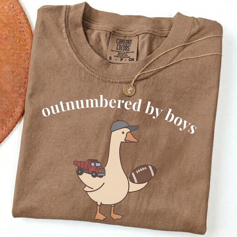 May include: A brown Comfort Colors t-shirt with the phrase "outnumbered by boys" in white. The shirt features a cartoon goose wearing a baseball cap and holding a football and a red truck. A gold necklace is draped across the shirt.