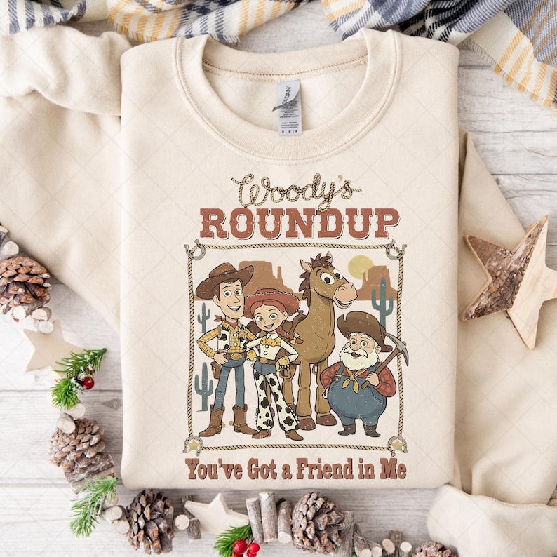 Op de afbeelding: Een cr&egrave;mekleurige sweatshirt met een afbeelding van Woody, Jessie, Bullseye en Stinky Pete uit Toy Story, met de tekst "Woody's Roundup" en "You've Got a Friend in Me". Het ontwerp is omlijst met een touwrand.