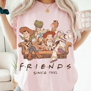 May include: Pink t-shirt featuring a graphic of characters from the Toy Story movie franchise. The design includes Woody, Buzz Lightyear, and other characters seated on a couch. The word "FRIENDS" is written below the graphic, with "SINCE 1995" underneath.