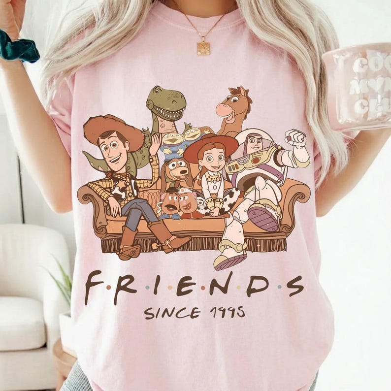 May include: Pink t-shirt featuring a graphic of characters from the Toy Story movie franchise. The design includes Woody, Buzz Lightyear, and other characters seated on a couch. The word "FRIENDS" is written below the graphic, with "SINCE 1995" underneath.