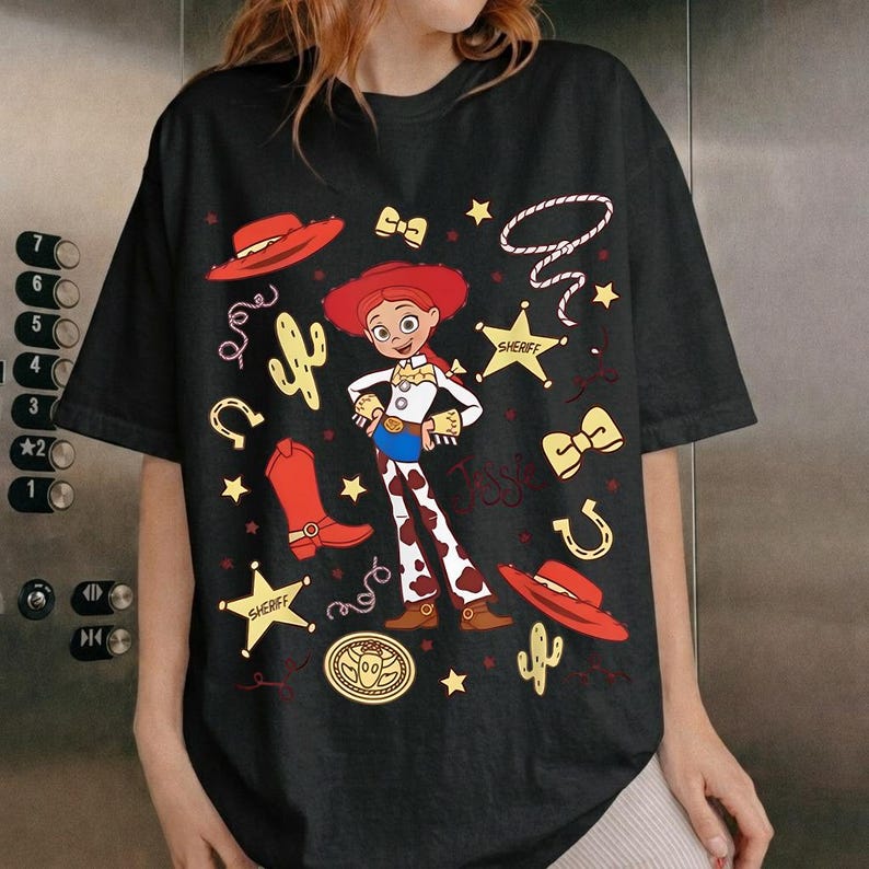 Puede incluir: Camiseta negra con una ilustraci&oacute;n de dibujos animados de una vaquera, que incluye sombrero rojo, botas y estrellas de sheriff. El dise&ntilde;o incluye el nombre del personaje y elementos tem&aacute;ticos del oeste.