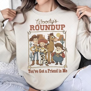 Op de afbeelding: Cr&egrave;mekleurige sweatshirt met een afbeelding van Woody, Jessie, Bullseye en Stinky Pete uit Toy Story. Het ontwerp bevat de tekst "Woody's Roundup" en "You've Got a Friend in Me" in een western-stijl lettertype, omlijst door een touwrand.