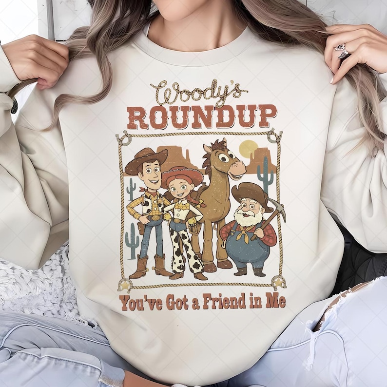 Op de afbeelding: Cr&egrave;mekleurige sweatshirt met een afbeelding van Woody, Jessie, Bullseye en Stinky Pete uit Toy Story. Het ontwerp bevat de tekst "Woody's Roundup" en "You've Got a Friend in Me" in een western-stijl lettertype, omlijst door een touwrand.