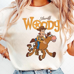 K&ouml;nnte beinhalten: Cremefarbenes T-Shirt mit einer Cartoon-Illustration von Sheriff Woody, der ein braunes Pferd reitet. Das Design enth&auml;lt das Wort "Woody" in orangefarbenen Blockbuchstaben mit einem Stern und das Wort "Sheriff" dar&uuml;ber.