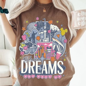 Puede incluir: Camiseta marr&oacute;n con un dise&ntilde;o gr&aacute;fico colorido que presenta im&aacute;genes ic&oacute;nicas de parques tem&aacute;ticos, incluyendo un castillo y un monorra&iacute;l, adem&aacute;s de flores estilizadas. El texto "What Dreams Are Made Of" se muestra.