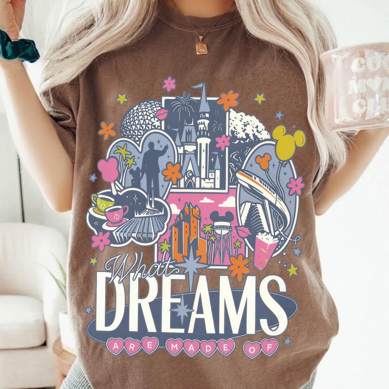 Puede incluir: Camiseta marr&oacute;n con un dise&ntilde;o gr&aacute;fico colorido que presenta im&aacute;genes ic&oacute;nicas de parques tem&aacute;ticos, incluyendo un castillo y un monorra&iacute;l, adem&aacute;s de flores estilizadas. El texto "What Dreams Are Made Of" se muestra.