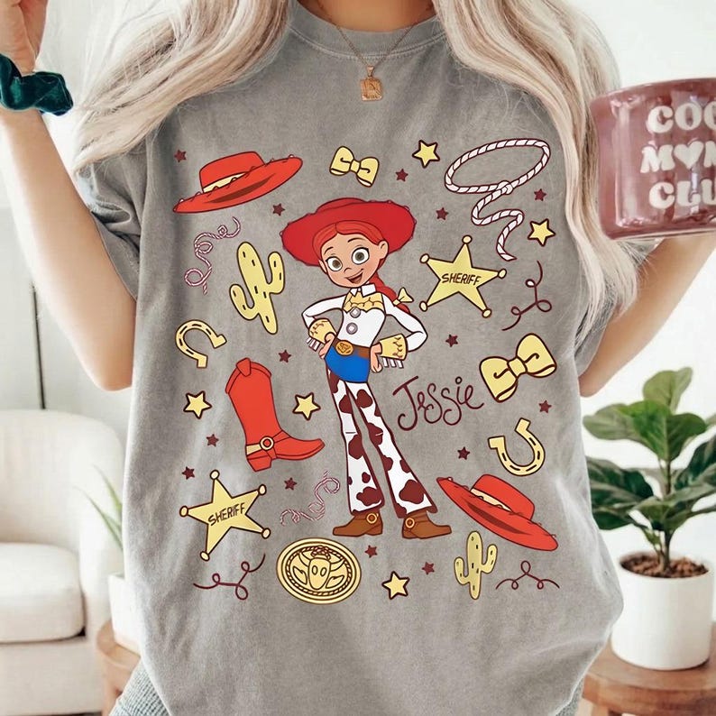 Puede incluir: Camiseta gris con una ilustraci&oacute;n de dibujos animados de Jessie de Toy Story. El dise&ntilde;o incluye un sombrero rojo, botas y la palabra "Jessie". Otros elementos incluyen estrellas, un cactus y una placa de sheriff.