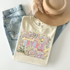 May include: Cream-colored t-shirt with a colorful graphic of Universal Studios attractions, including the text "UNIVERSAL STUDIOS." The shirt is paired with light-wash denim jeans, a gold necklace, and a tan hat. A sprig of dried flowers adds a decorative touch.