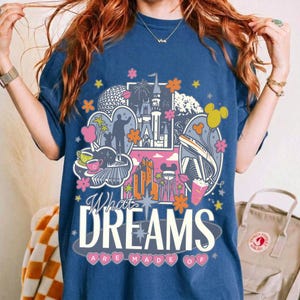Puede incluir: Camiseta azul marino con un dise&ntilde;o gr&aacute;fico colorido que presenta monumentos y personajes ic&oacute;nicos. El texto "What Dreams Are Made Of" se muestra en una fuente estilizada. Se incorporan acentos rosas, naranjas y amarillos.
