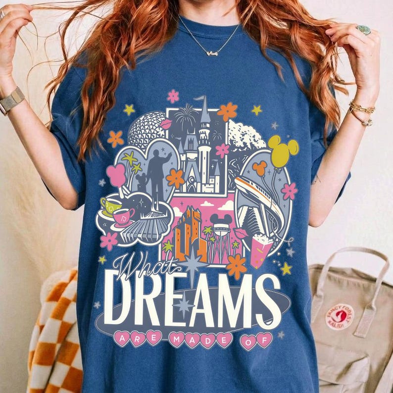 Puede incluir: Camiseta azul marino con un dise&ntilde;o gr&aacute;fico colorido que presenta monumentos y personajes ic&oacute;nicos. El texto "What Dreams Are Made Of" se muestra en una fuente estilizada. Se incorporan acentos rosas, naranjas y amarillos.