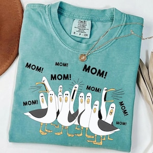 May include: A teal Comfort Colors t-shirt featuring a graphic of seagulls with the word "MOM!" repeated around them. A gold necklace with a pendant is draped across the shirt. The shirt is folded and has a tag.
