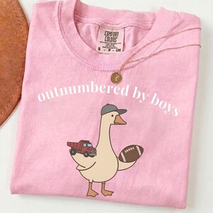 May include: Pink t-shirt with the text "outnumbered by boys" and a cartoon goose wearing a baseball cap, holding a football and a red truck. A gold necklace with a round pendant is on the shirt.