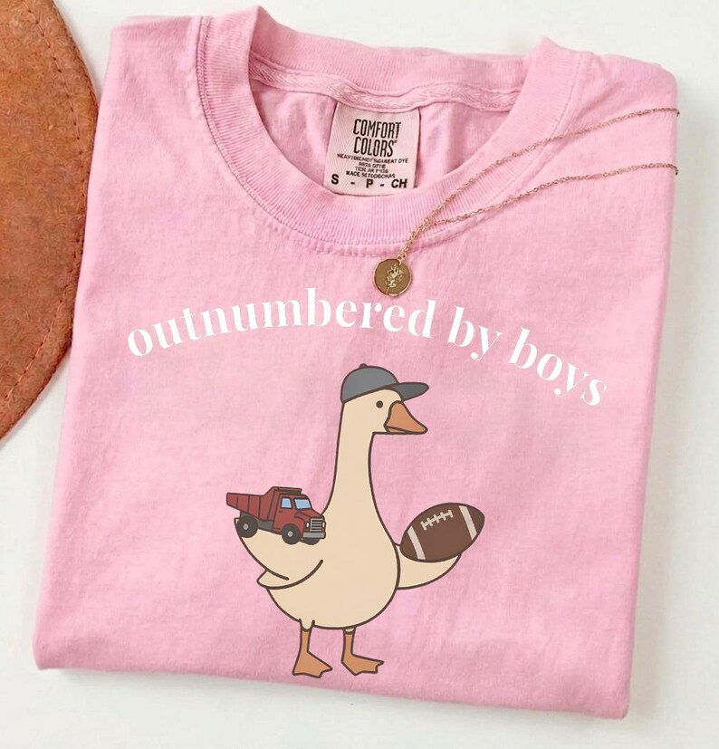 May include: Pink t-shirt with the text "outnumbered by boys" and a cartoon goose wearing a baseball cap, holding a football and a red truck. A gold necklace with a round pendant is on the shirt.