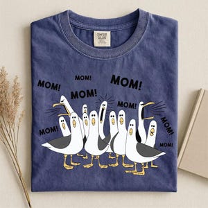 Pu&ograve; includere: T-shirt blu navy Comfort Colors con un'illustrazione a cartoni animati di gabbiani che gridano la parola "MOM!". I gabbiani sono bianchi con ali grigie e zampe gialle. La maglietta ha un girocollo.