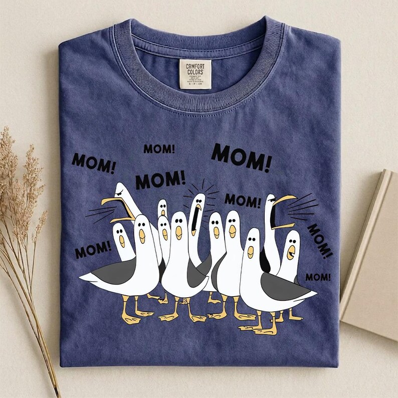 Pu&ograve; includere: T-shirt blu navy Comfort Colors con un'illustrazione a cartoni animati di gabbiani che gridano la parola "MOM!". I gabbiani sono bianchi con ali grigie e zampe gialle. La maglietta ha un girocollo.