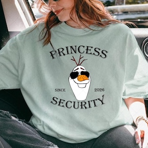 以下が含まれることがあります： 黒い文字で「PRINCESS SECURITY」と書かれた、サングラスとニンジンの鼻をした漫画の雪だるまが特徴の、明るい緑色のスウェットシャツ。「SINCE 2026」の文字も含まれています。スウェットシャツはカジュアルなスタイルです。