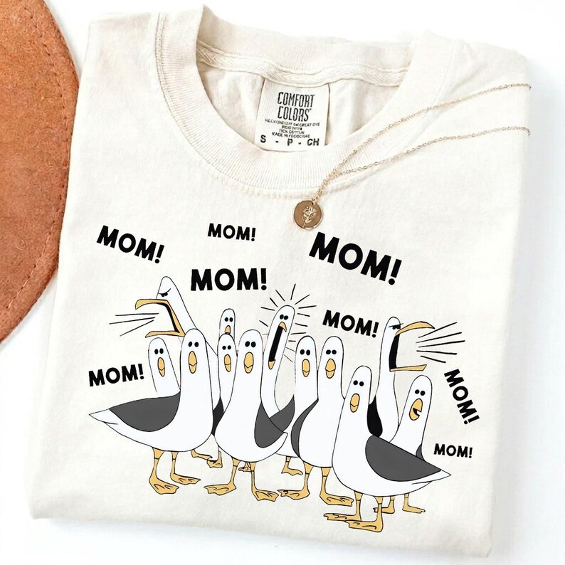 Pu&ograve; includere: T-shirt color crema con una grafica di gabbiani dei cartoni animati e la parola "MOM!" ripetuta. La maglietta ha un girocollo e maniche corte. Una collana d'oro con un ciondolo &egrave; drappeggiata sulla maglietta.