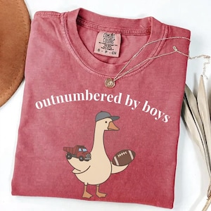 May include: A red Comfort Colors t-shirt with the phrase "outnumbered by boys" in white. The shirt features a cartoon goose wearing a baseball cap, carrying a toy truck and a football. A gold necklace is draped on the shirt.