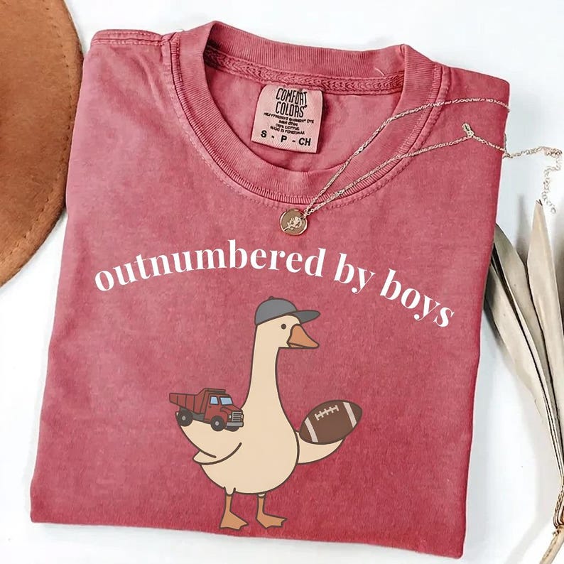 May include: A red Comfort Colors t-shirt with the phrase "outnumbered by boys" in white. The shirt features a cartoon goose wearing a baseball cap, carrying a toy truck and a football. A gold necklace is draped on the shirt.