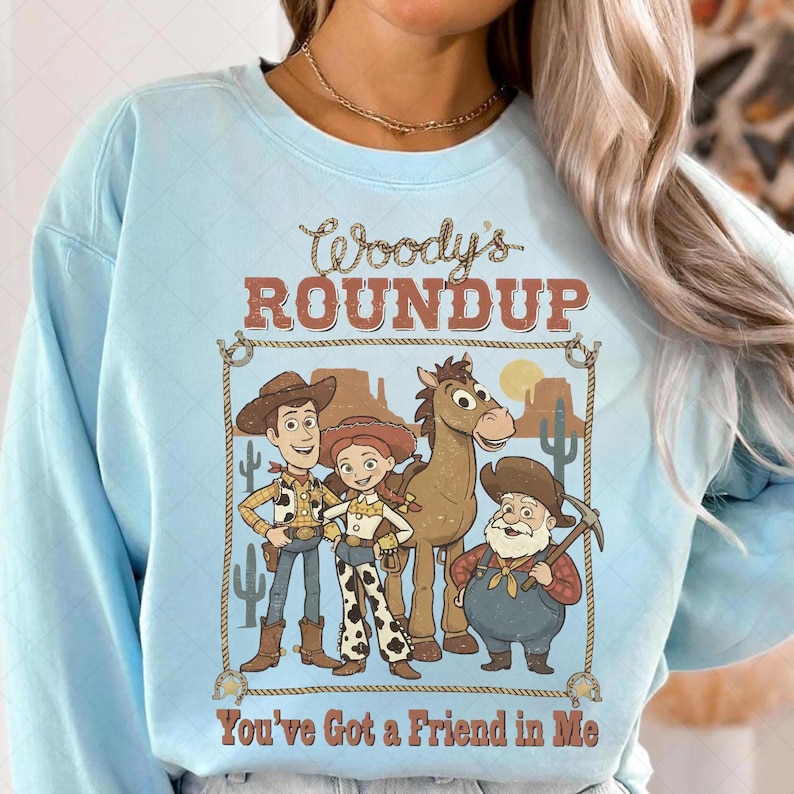 Op de afbeelding: Lichtblauw sweatshirt met een afbeelding van Woody, Jessie, Bullseye en Stinky Pete uit Toy Story. Het ontwerp bevat de tekst "Woody's Roundup" en "You've Got a Friend in Me" in een western-stijl lettertype.