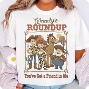 Op de afbeelding: Wit t-shirt met een cartoonafbeelding van personages uit een animatiefilm. Het ontwerp bevat Woody, Jessie en Bullseye, met de tekst "Woody's Roundup" en "You've Got a Friend in Me" in een bruin lettertype.