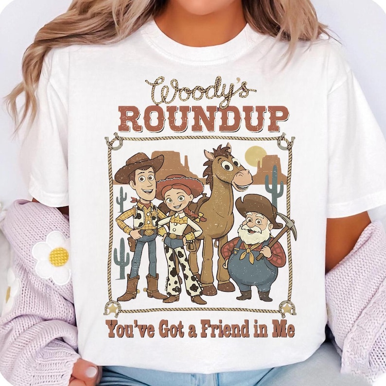 Op de afbeelding: Wit t-shirt met een cartoonafbeelding van personages uit een animatiefilm. Het ontwerp bevat Woody, Jessie en Bullseye, met de tekst "Woody's Roundup" en "You've Got a Friend in Me" in een bruin lettertype.