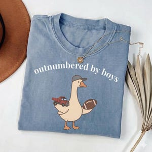 May include: A blue t-shirt with the phrase "outnumbered by boys" in white. The shirt features a cartoon goose wearing a baseball cap and holding a football and a red truck. A gold necklace is draped on the shirt.