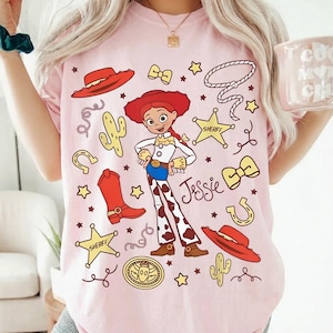 Puede incluir: Camiseta rosa claro con un personaje de dibujos animados de vaquera llamado Jessie, rodeado de ilustraciones tem&aacute;ticas de vaqueros. El dise&ntilde;o incluye un sombrero rojo, botas, insignias de sheriff y la palabra "Jessie".