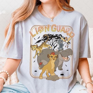 Könnte beinhalten: Hellblaues T-Shirt mit einem farbenfrohen Grafikdesign der Charaktere der Lion Guard. Das Design zeigt ein Löwenjunges, einen Geparden, ein Nilpferd, einen Vogel und ein graues Tier, mit dem Schriftzug "Lion Guard" in Gold und Orange.