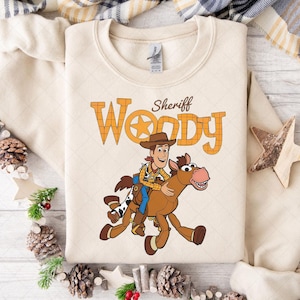 K&ouml;nnte beinhalten: Cremefarbenes Sweatshirt mit einer Grafik von Sheriff Woody auf einem Pferd. Der Text "Sheriff Woody" ist oberhalb der Illustration in Orange und Braun aufgedruckt. Das Sweatshirt hat einen Rundhalsausschnitt und lange &Auml;rmel.