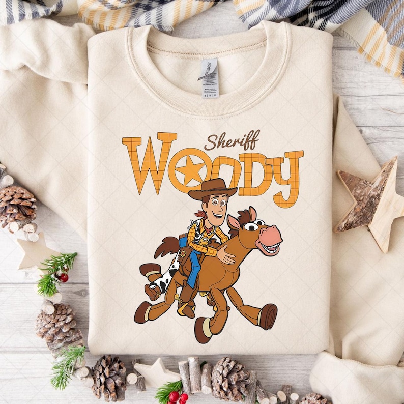 K&ouml;nnte beinhalten: Cremefarbenes Sweatshirt mit einer Grafik von Sheriff Woody auf einem Pferd. Der Text "Sheriff Woody" ist oberhalb der Illustration in Orange und Braun aufgedruckt. Das Sweatshirt hat einen Rundhalsausschnitt und lange &Auml;rmel.