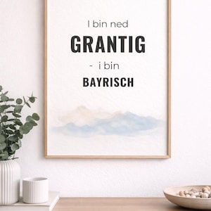 Könnte beinhalten: Holzgerahmter Druck mit dem Text "I bin ned GRANTIG - i bin BAYRISCH" in Schwarz, über einer Aquarell-Berglandschaft in Blau und Beige. Der Druck wird auf einem Holzschrank ausgestellt.