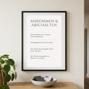 Op de afbeelding: Een ingelijste print met de Duitse tekst "ANKOMMEN & ABSCHALTEN". De zwarte lijst omringt een witte achtergrond met zwarte tekst. Een kleine kom met een telefoon en hoesje staat op een houten oppervlak eronder.