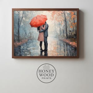Puede incluir: Una impresión artística enmarcada representa a una pareja abrazándose bajo un paraguas rojo en un bosque lluvioso. La obra de arte utiliza azules, grises y naranjas apagados, con reflejos en el suelo mojado. La impresión está enmarcada en madera oscura.