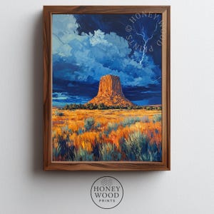 May include: A framed art print depicting a large, orange-hued rock formation rising from a golden-yellow grassy plain under a dark, stormy blue sky with a visible lightning bolt. The wooden frame surrounds the rectangular landscape painting.