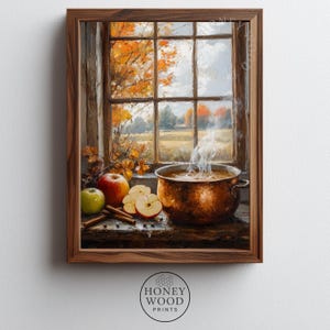 May include: A framed wall art print depicting a copper pot filled with steaming liquid, placed on a wooden surface beside sliced apples, a whole green apple, a red apple, and cinnamon sticks, with a window view of autumn trees.