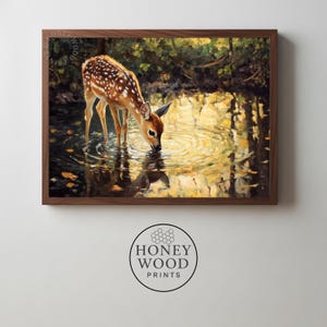 May include: A framed art print depicts a young deer drinking from a pond. The deer has brown fur with white spots. The water reflects the deer and the surrounding trees. The artwork is in a wooden frame. The print is from Honey Wood Prints.