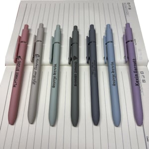 Op de afbeelding: Zeven kleurrijke pennen met zwarte clips zijn gerangschikt op een notitieblok. De pennen zijn in tinten roze, grijs, blauwgroen, donkergrijs, blauwgrijs, lichtblauw en lavendel. Elke pen heeft tekst erop, waaronder bedrijfsnamen en persoonsnamen.
