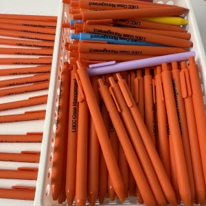 Op de afbeelding: Een verzameling oranje pennen met zwarte tekst "LHCC Case Management" gerangschikt in een witte plastic container. Een paar pennen in andere kleuren, waaronder blauw, geel en paars, zijn ook zichtbaar.