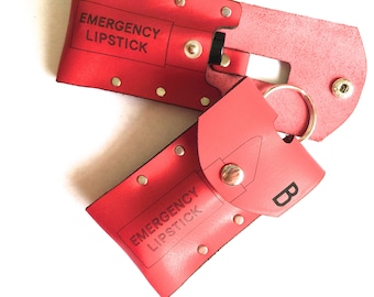 May include: A red leather keychain with a lipstick holder and a key ring. The keychain has the text "EMERGENCY LIPSTICK" on it and the letter "B" on the other side.