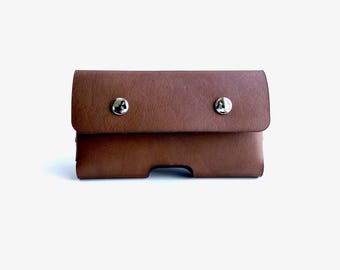 May include: A brown leather phone case with a flap closure secured by two silver snap buttons. The case has a rectangular shape with a cutout at the bottom. The leather has a textured appearance.