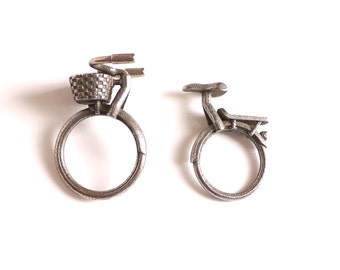 May include: Two silver metal rings shaped like bicycles. The ring on the left has a basket on the front. The ring on the right has a seat and handlebars.