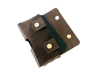 May include: A brown leather phone case with a magnetic closure. The case is open, revealing a black phone inside. The case has a textured finish and gold-colored snap closures.