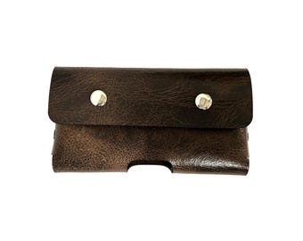 May include: A dark brown leather phone case with a flap closure secured by two silver snap buttons. The case has a textured appearance and a cutout for the phone's features. The overall design is simple and functional.