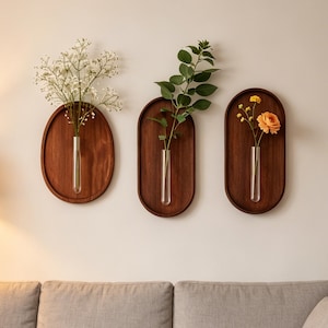May include: Three wooden wall vases with glass tubes holding flowers and greenery. The vases are oval-shaped and have a dark brown finish. The flowers include white, green, and orange blooms, creating a natural and decorative display.