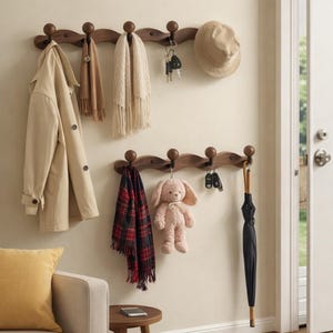 Puede incluir: Dos percheros de madera con múltiples ganchos están montados en una pared beige. Un abrigo color canela, bufandas, llaves y un sombrero cuelgan del perchero superior. El perchero inferior sostiene una bufanda a cuadros, un conejito de peluche, llaves y un paraguas negro.