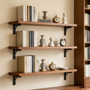 May include: Three wooden shelves with black metal brackets are mounted on a beige wall. The shelves display books, small clocks, and decorative figurines. The shelves are made of dark wood and are spaced evenly apart.