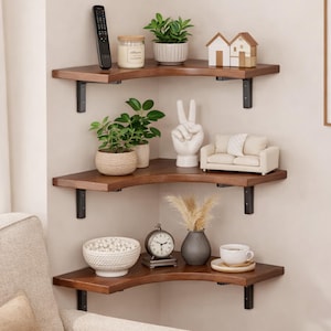 Puede incluir: Estanterías esquineras de tres niveles hechas de madera oscura, cada una soportada por soportes metálicos negros. Las estanterías están decoradas con pequeñas plantas en macetas, objetos decorativos, un mando a distancia y una vela.