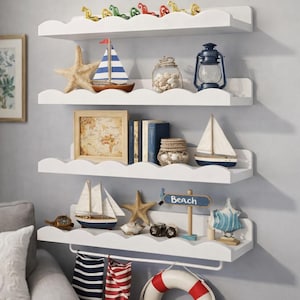 Puede incluir: Estanterías blancas en forma de nube exhiben decoración náutica. Incluye maquetas de veleros, estrellas de mar, una linterna, un mapa enmarcado, libros y tarros decorativos. Un letrero dice "Beach". Las estanterías están montadas en una pared gris claro.