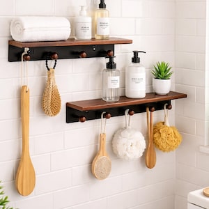 Könnte beinhalten: Zweistöckiges Badezimmerregal mit schwarzen Metallhalterungen, dunklen Holzregalen und Holzknöpfen. Die Regale beherbergen ein weißes Handtuch, Seifenflaschen und eine kleine Topfpflanze. Verschiedene Badezimmeraccessoires hängen an den Knöpfen.